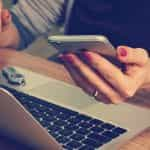 A woman sitting in front of her laptop at a desk while holding a smartphone in her left hand.