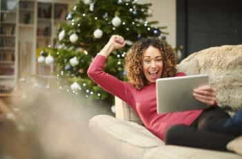 A smiling and happy lady celebrates a winner while watching an event on her tablet led on the sofa.