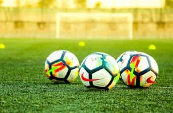 Three soccer balls sit on a green field with a net in the distance behind them.