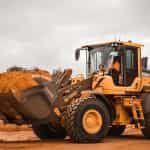 A yellow bulldozer pauses in the midst of reddish sand, driven by someone in an orange uniform.