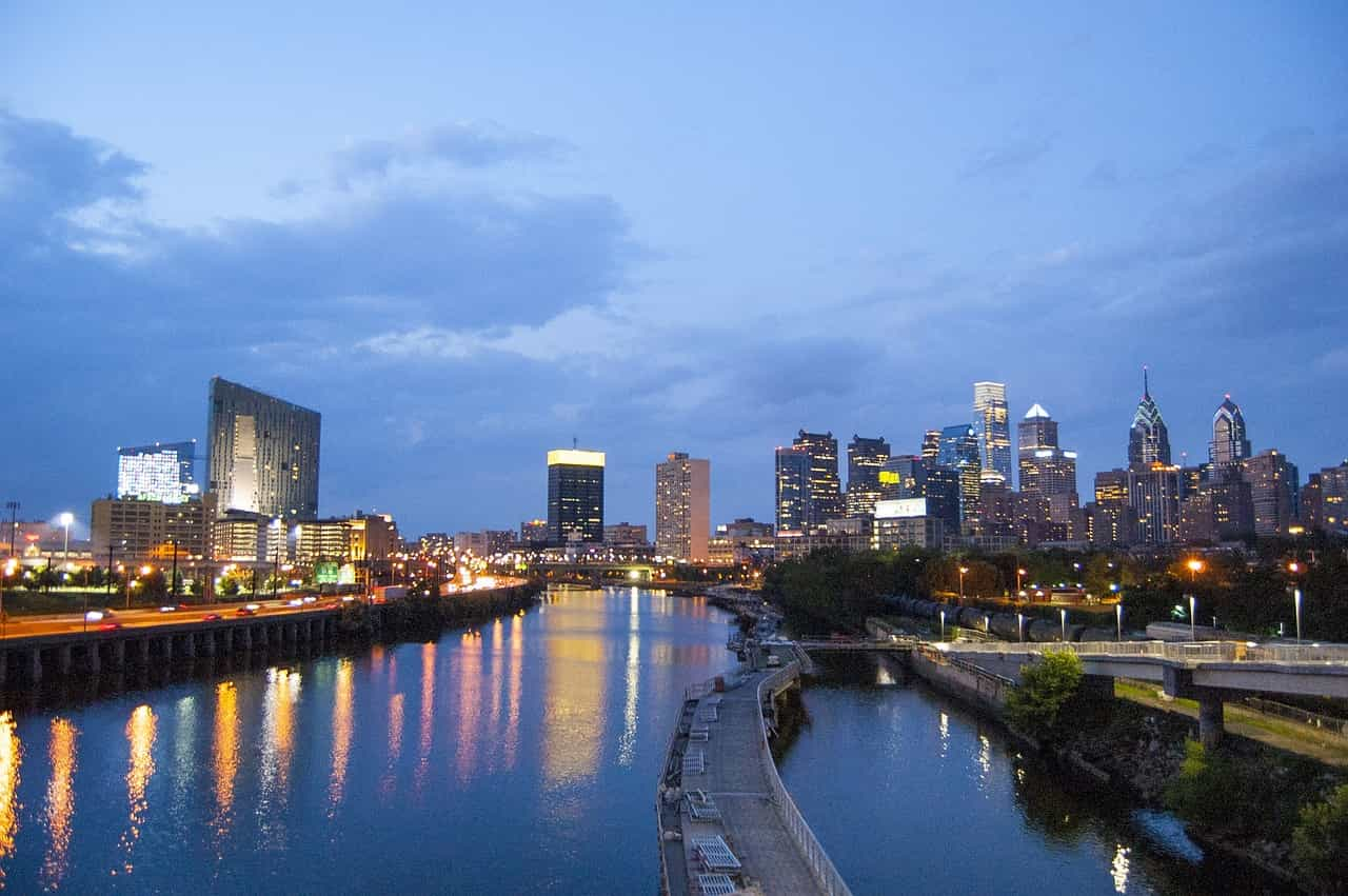 A view of downtown Philadelphia downriver from the city, with a long body of water diving two sides of the city from one another, with the city’s dense downtown area full of tall building and skyscrapers to the right. 