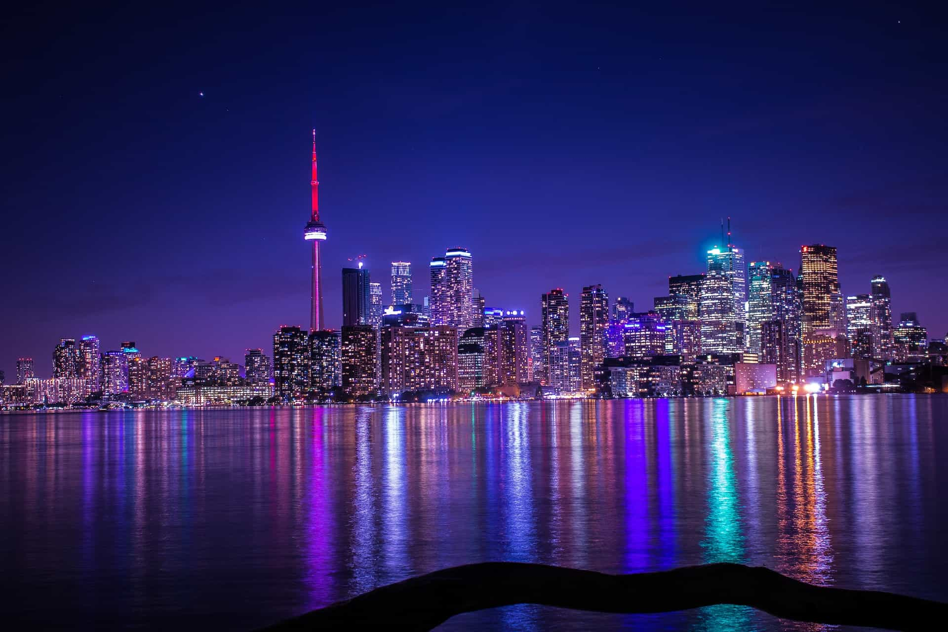 The bustling skyline of downtown Toronto, Ontario in Canada, featuring dozens of illuminated skyscrapers and tall buildings and the iconic CN Tower, with Lake Ontario stretching out in front of it.