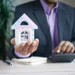 A man in a suit holding a small model house in the palm of his hand.